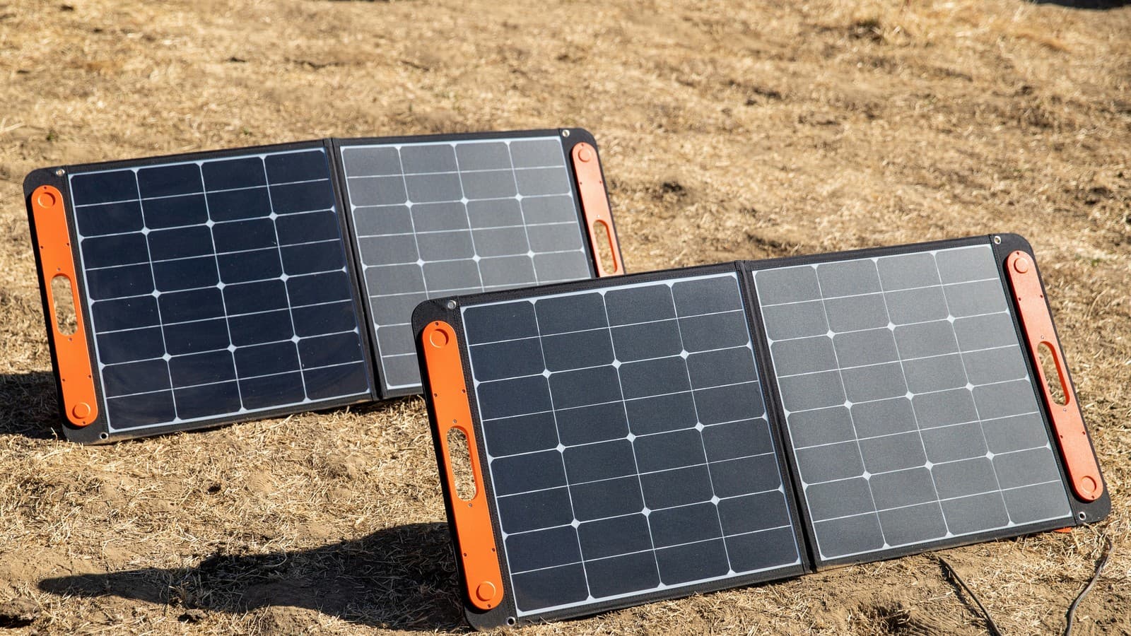 Two unfolded portable solar panels with orange carry handles standing on dry ground.
