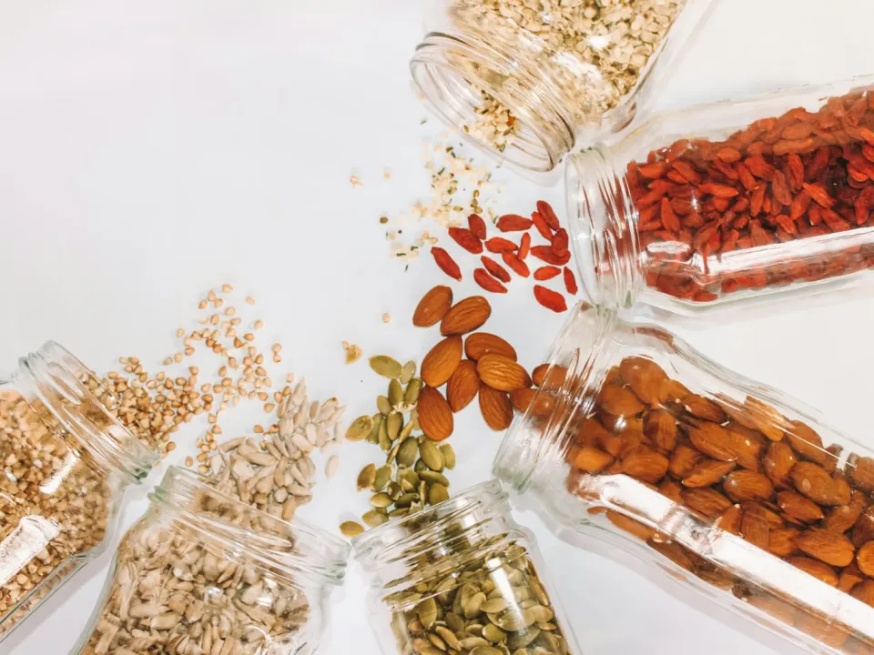 Open glass jars of almonds, pumpkin seeds, sunflower seeds, buckwheat and dried goji berries spilling onto a white table